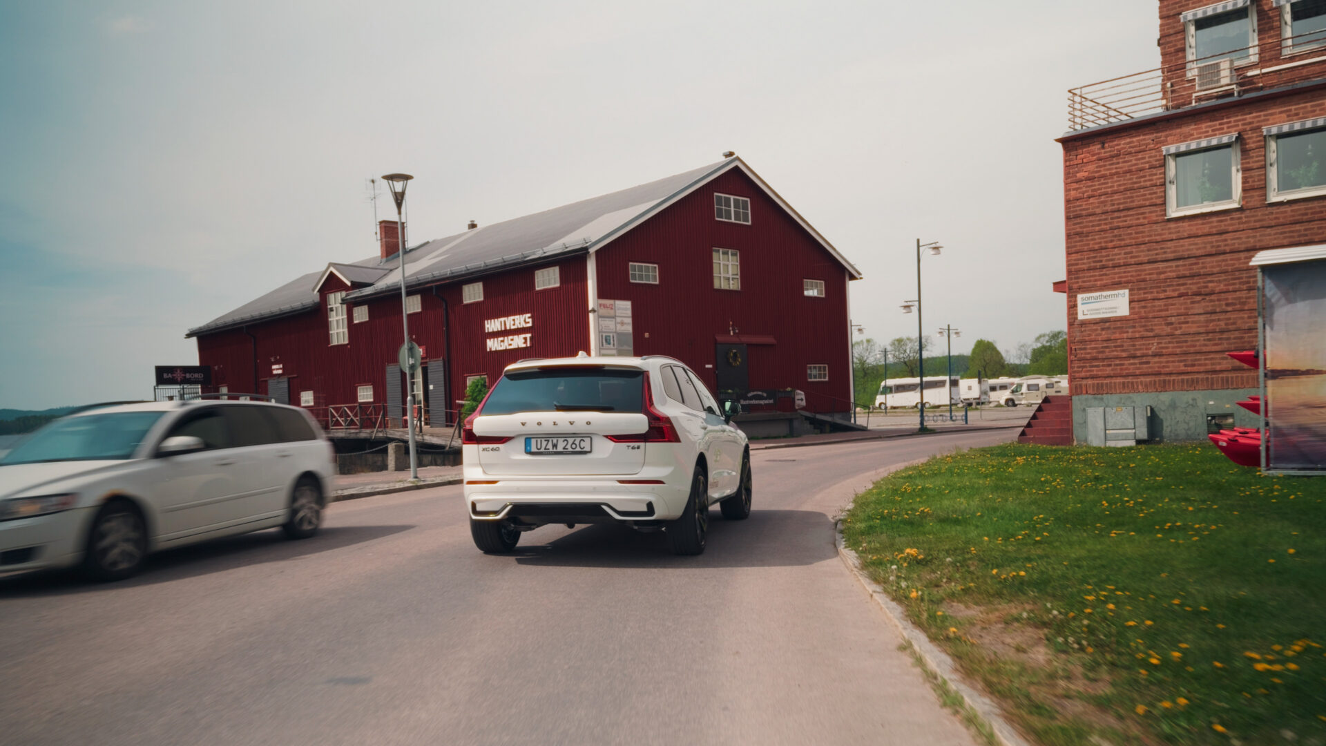 Vit Volvo XC60 kör på gata förbi rött trähus och träbyggnad i Arvika under diffust dagsljus i Helmia