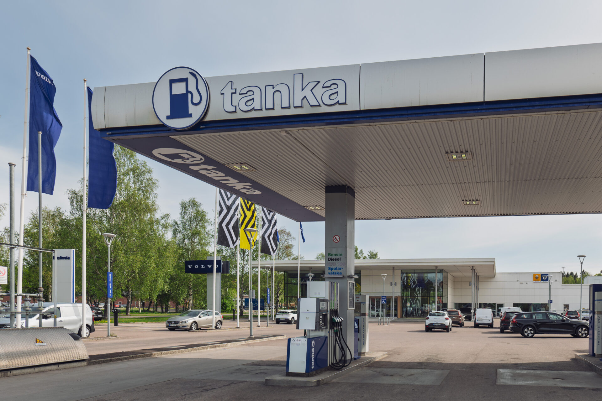 Tanka bensinstation med flera bränslepumpar och Volvo-flaggor vid Helmia bilhall i Arvika under en mild molnig dag