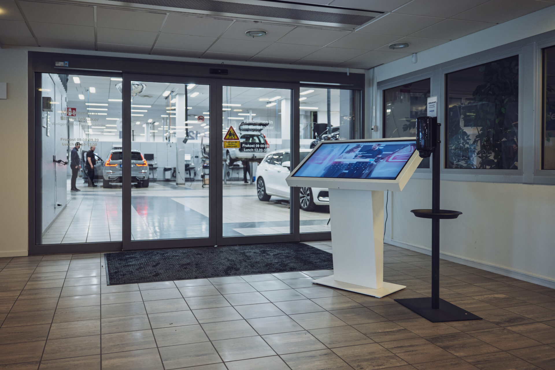 Interiör på Helmia bilverkstad i Karlstad med en digital informationskiosk och automatiska glasdörrar där Volvo-bilar och personal syns i verkstadsdelen