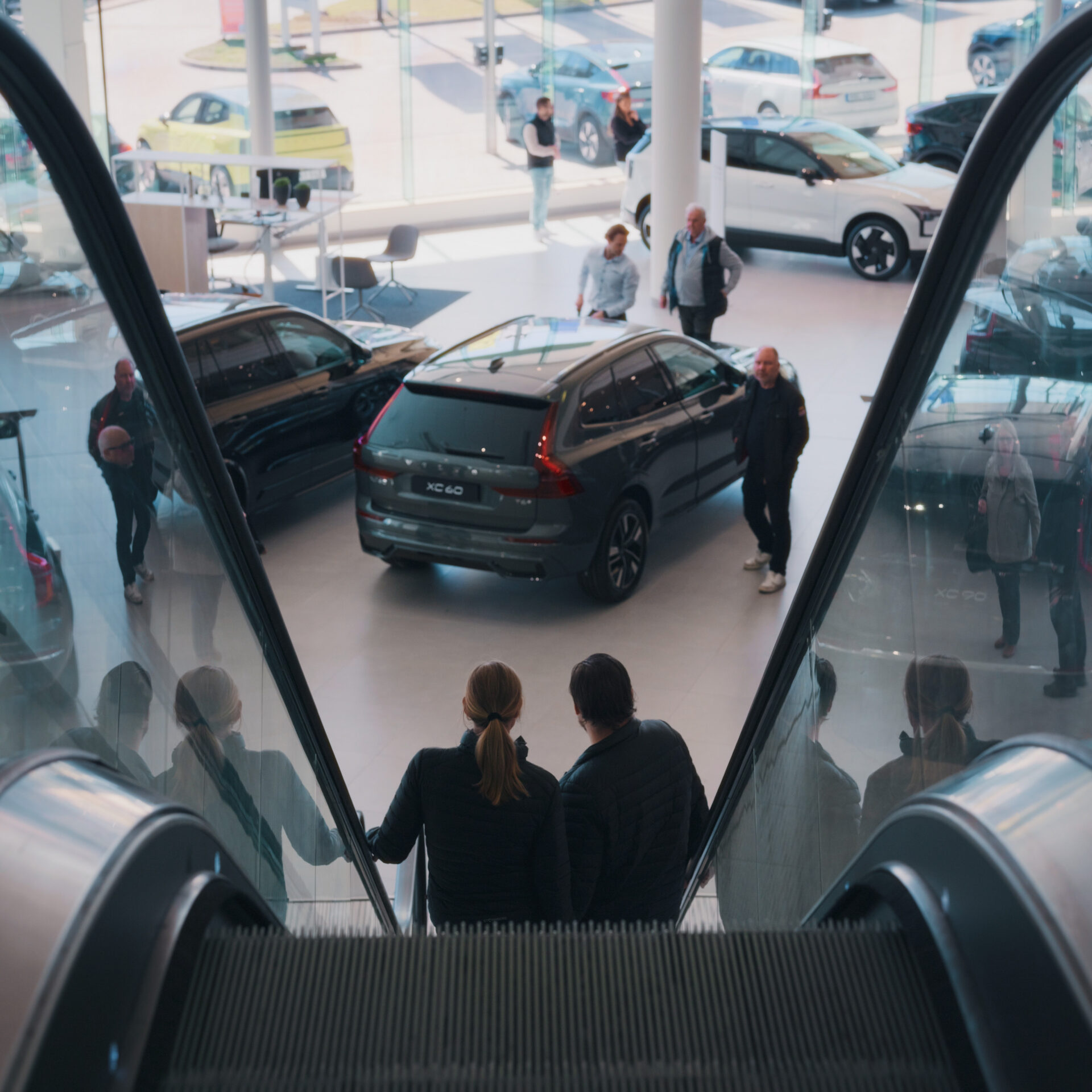Par som åker rulltrappa ner mot en rymlig bilhall med Volvo-bilar och personer som tittar på fordonen hos Helmia i Karlstad