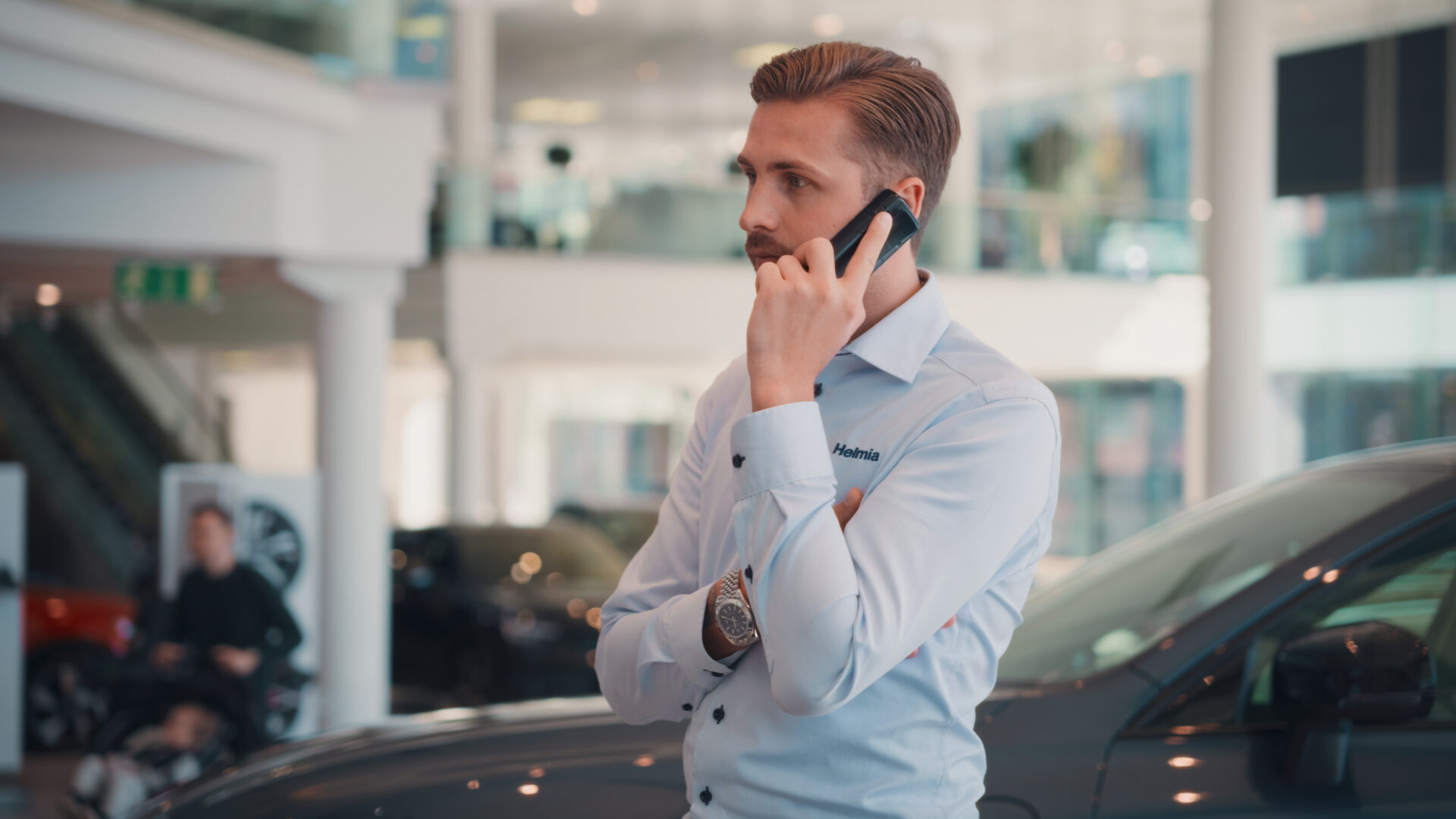 Ung man i ljusblå skjorta med Helmia-logotyp pratar i mobiltelefon inne i modern bilhall i Karlstad i Volvo
