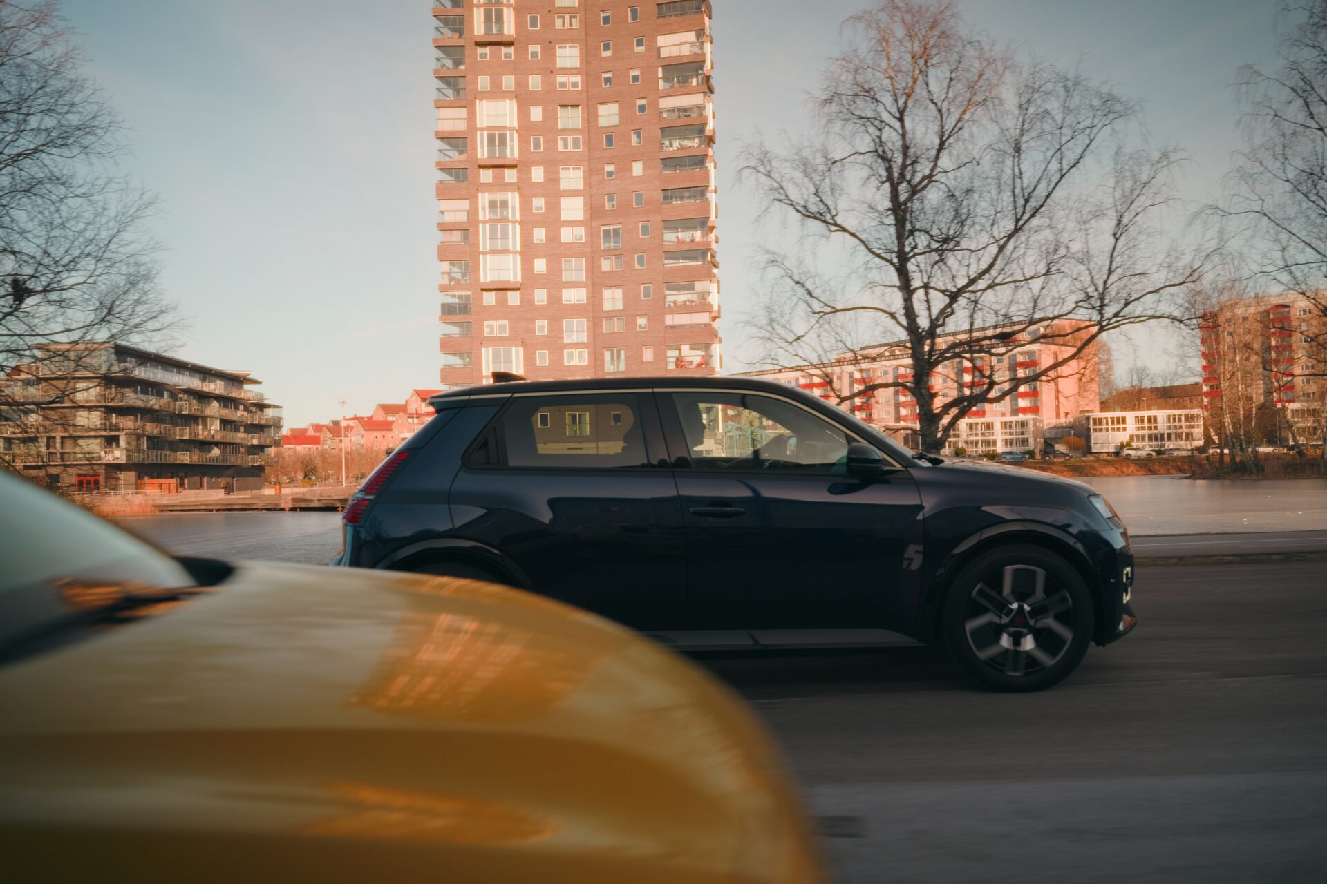 Mörk Renault 5 i sidoprofil körande genom Karlstads stad med moderna bostadshus och kala träd i varmt gyllene solljus i Helmia