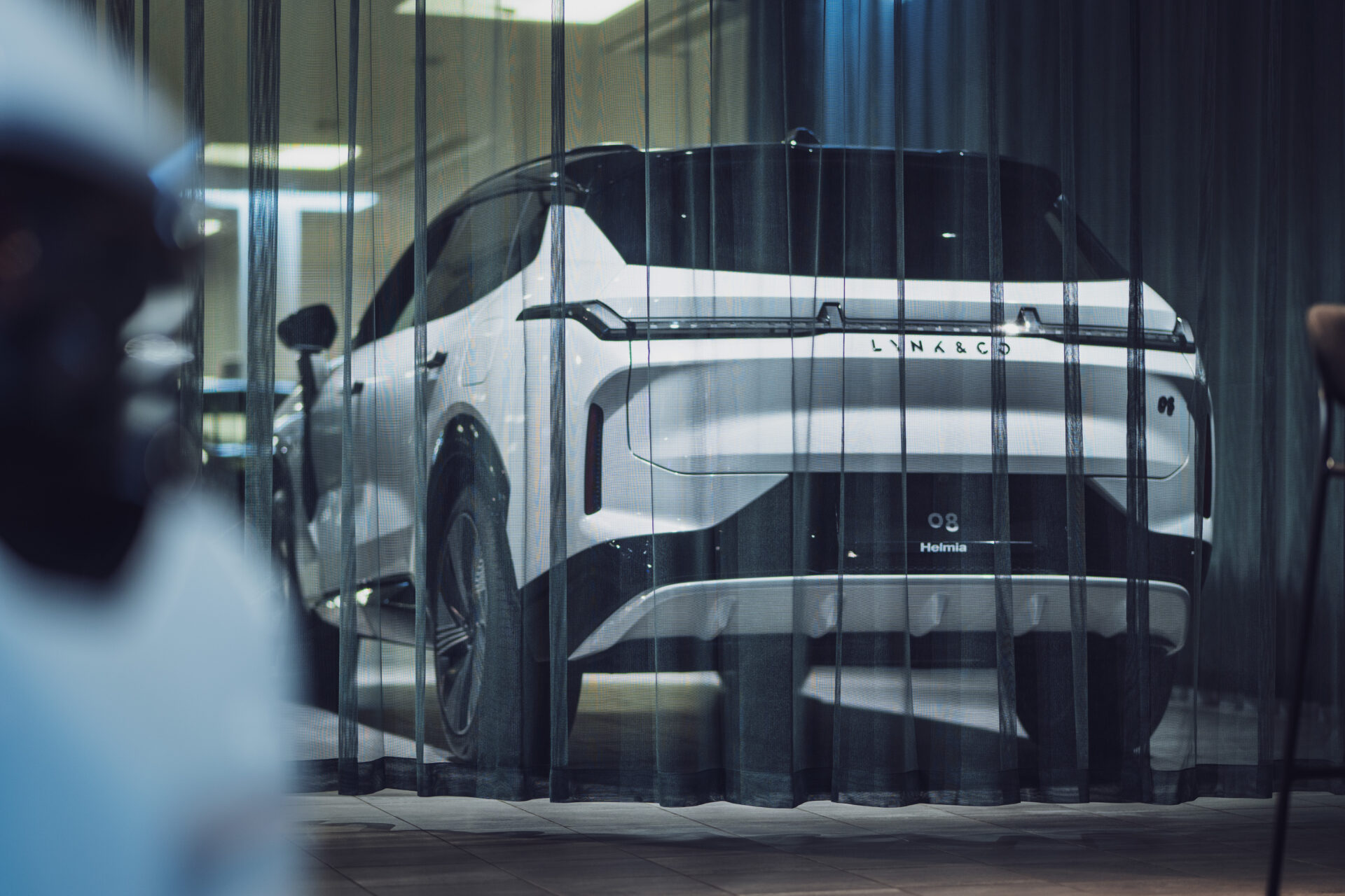 Vit Lynk & Co Helmia 08 el-SUV sedd genom svart genomskinligt draperi i en mjukt belyst inomhusmiljö