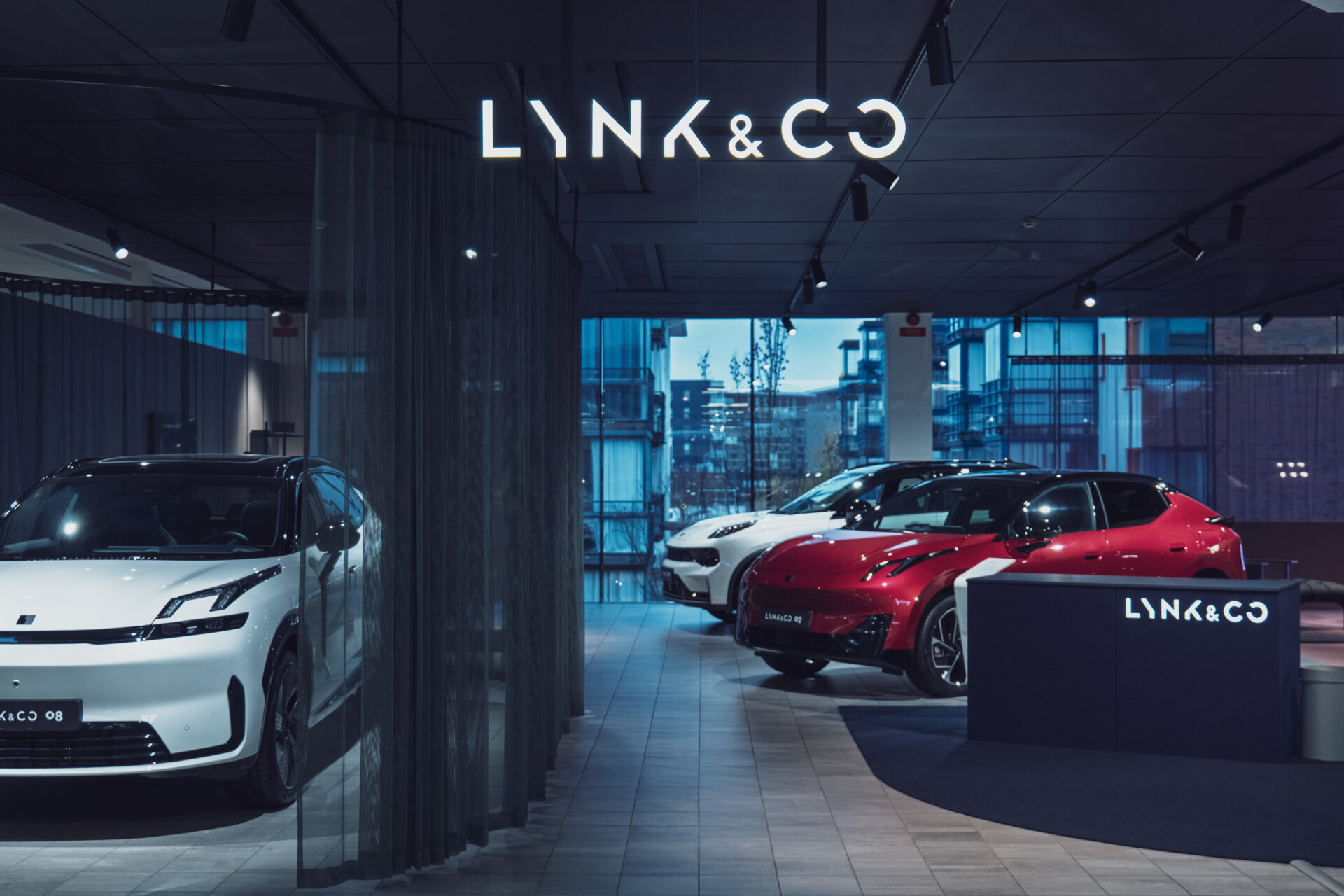 Interiör av en modern Helmia Lynk & Co bilhall i Lynk med vita och röda elbilar belysta av mjukt takljus och stadsbyggnader synliga genom stora fönster