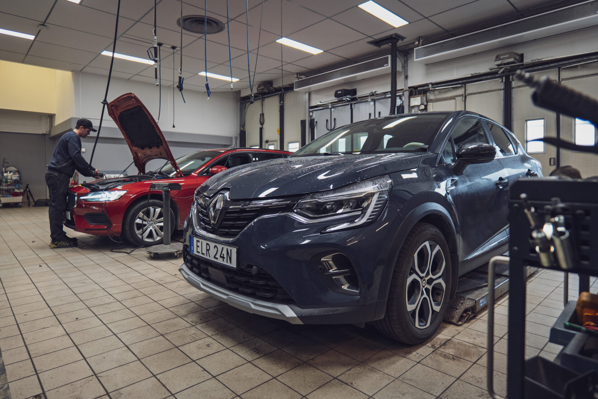 Helmia bilverkstad i Sunne med två bilar, en mörk Renault SUV och en röd bil med öppen motorhuv, och en mekaniker som arbetar på den röda bilen.