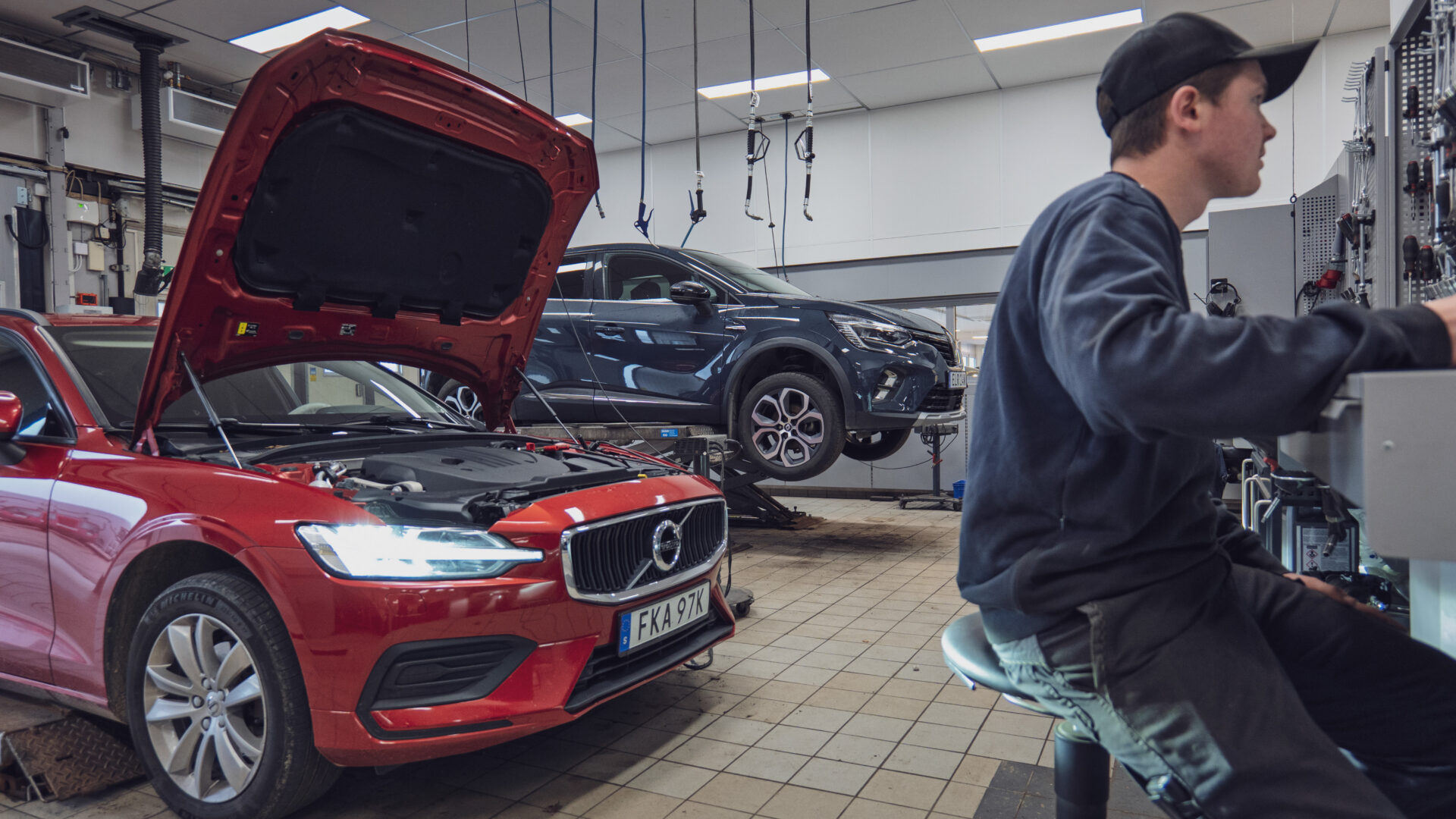 Interiör från Helmia bilverkstad i Värmland med mekaniker vid arbetsstation och två fordon, Volvo med öppen motorhuv och en annan bil på lyft i Sunne
