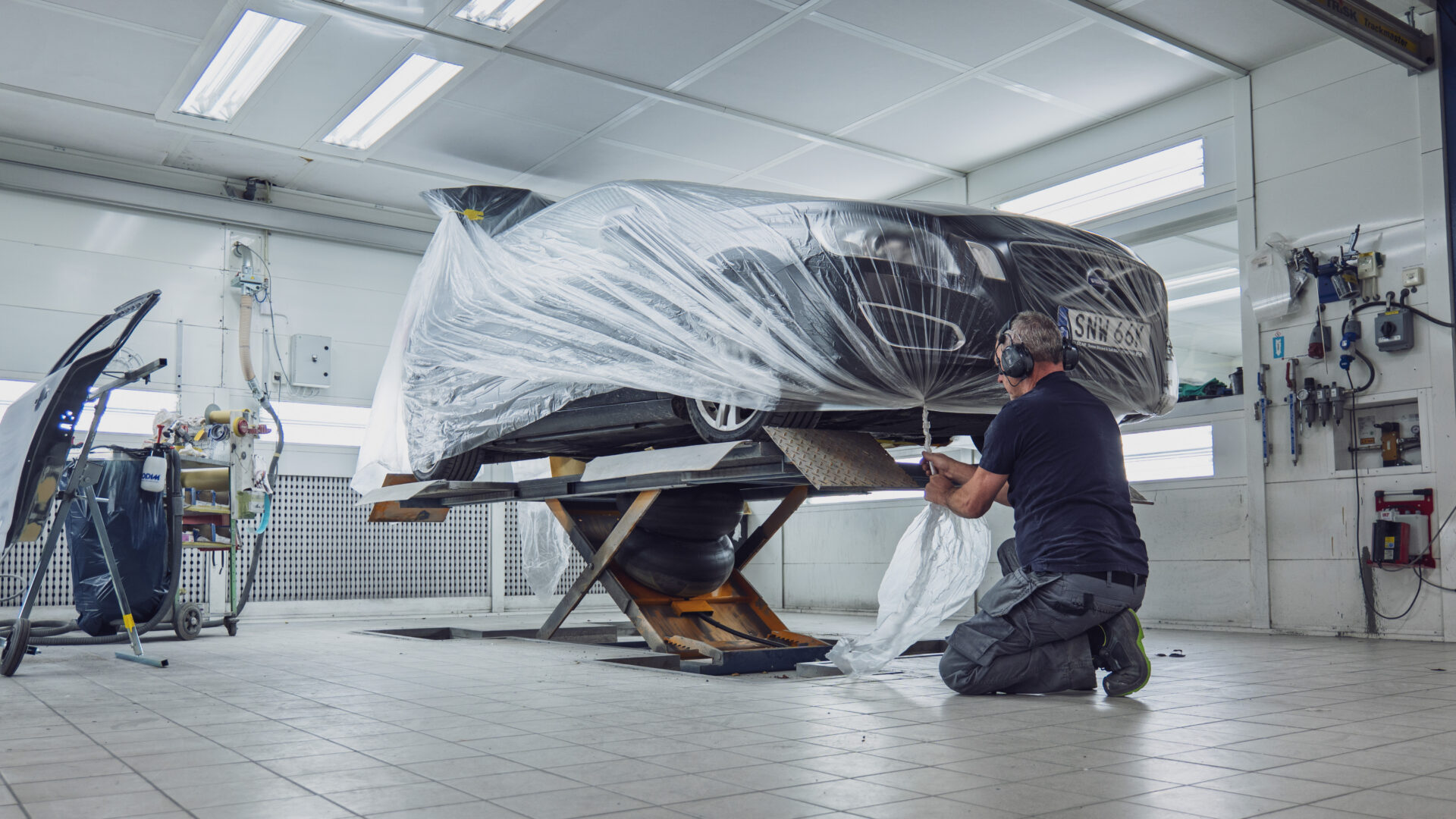 Helmia mekaniker knäböjer bredvid en Volvo bil täckt med skyddsplast på en hydraulisk lyft i en väl upplyst verkstad i Sunne