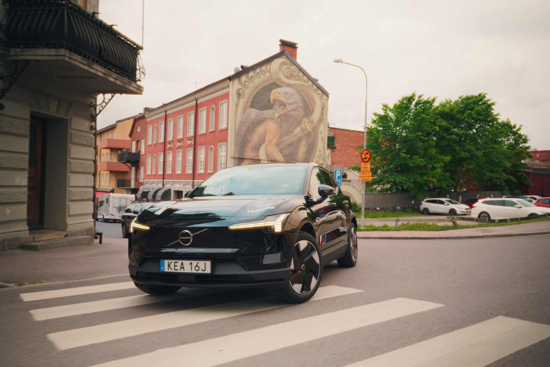 Helmia Volvo EX30 elbil i svart färg som korsar ett övergångsställe i Arvika med en stor örnmålning på en byggnad i bakgrunden