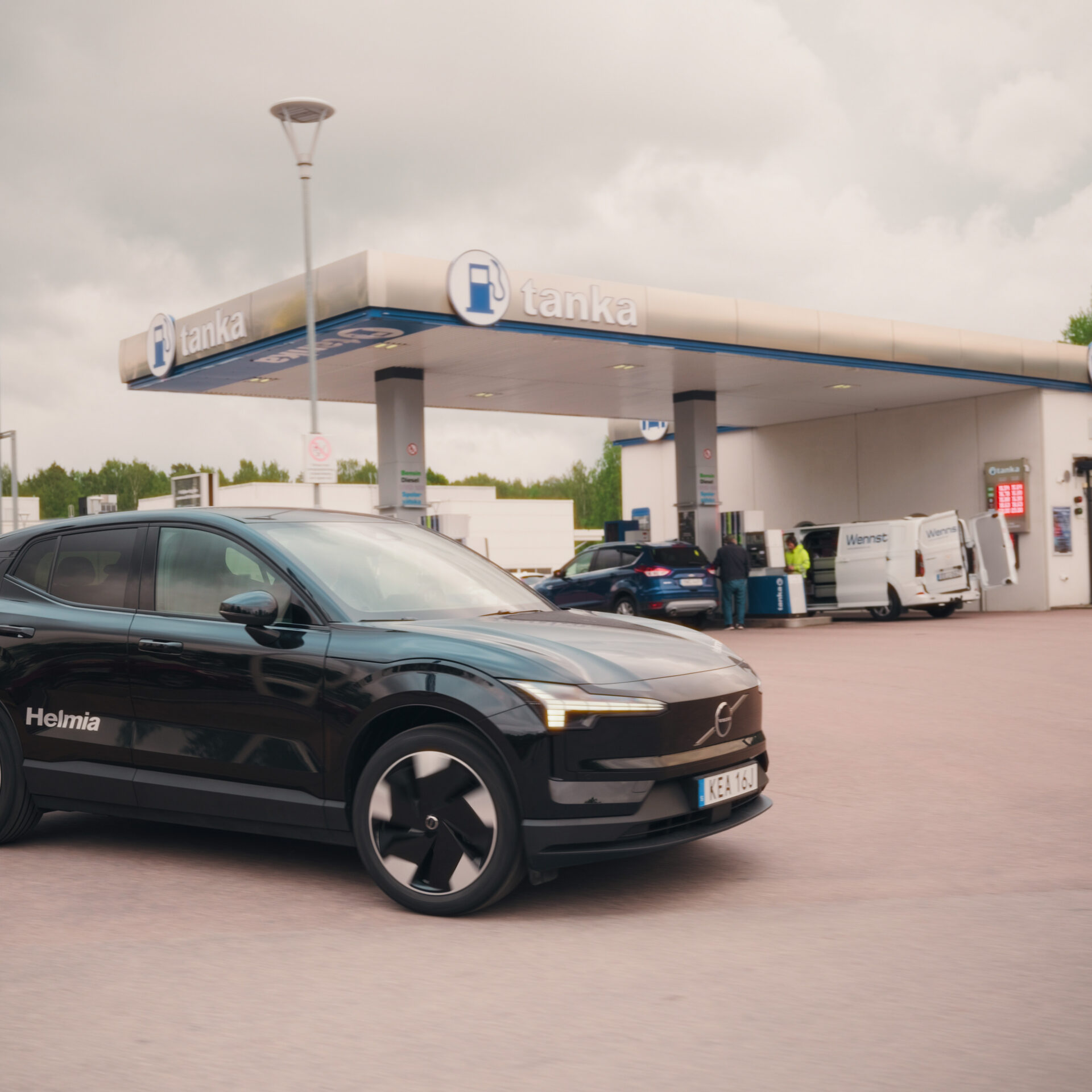 Modern svart Volvo EX30 el-SUV från Helmia parkerad vid en bensinstation i Arvika med grönska och molnig himmel i bakgrunden