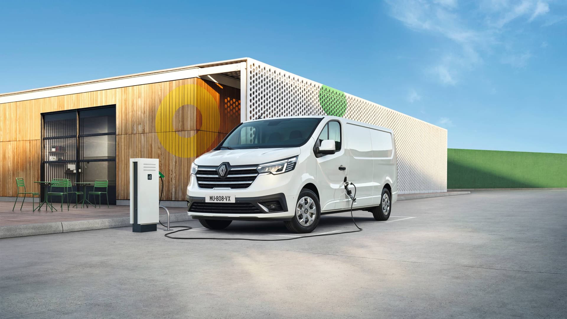Vit Renault Kangoo E-Tech electric skåpbil laddar vid en laddstation utanför en modern byggnad med träpaneler och vit perforerad fasad under klarblå himmel i Helmia