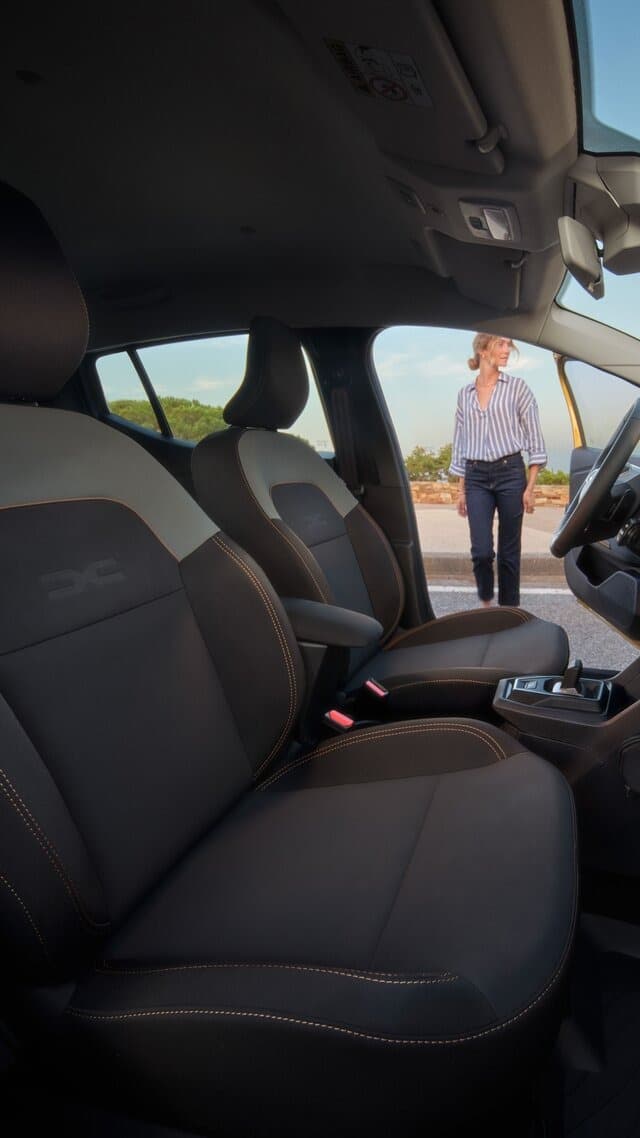 Interiörbild av Dacia Sandero Stepway med framstolar i svart och orange sömnad, förardörr öppen och kvinna stående utanför bilen i dagsljus i Helmia